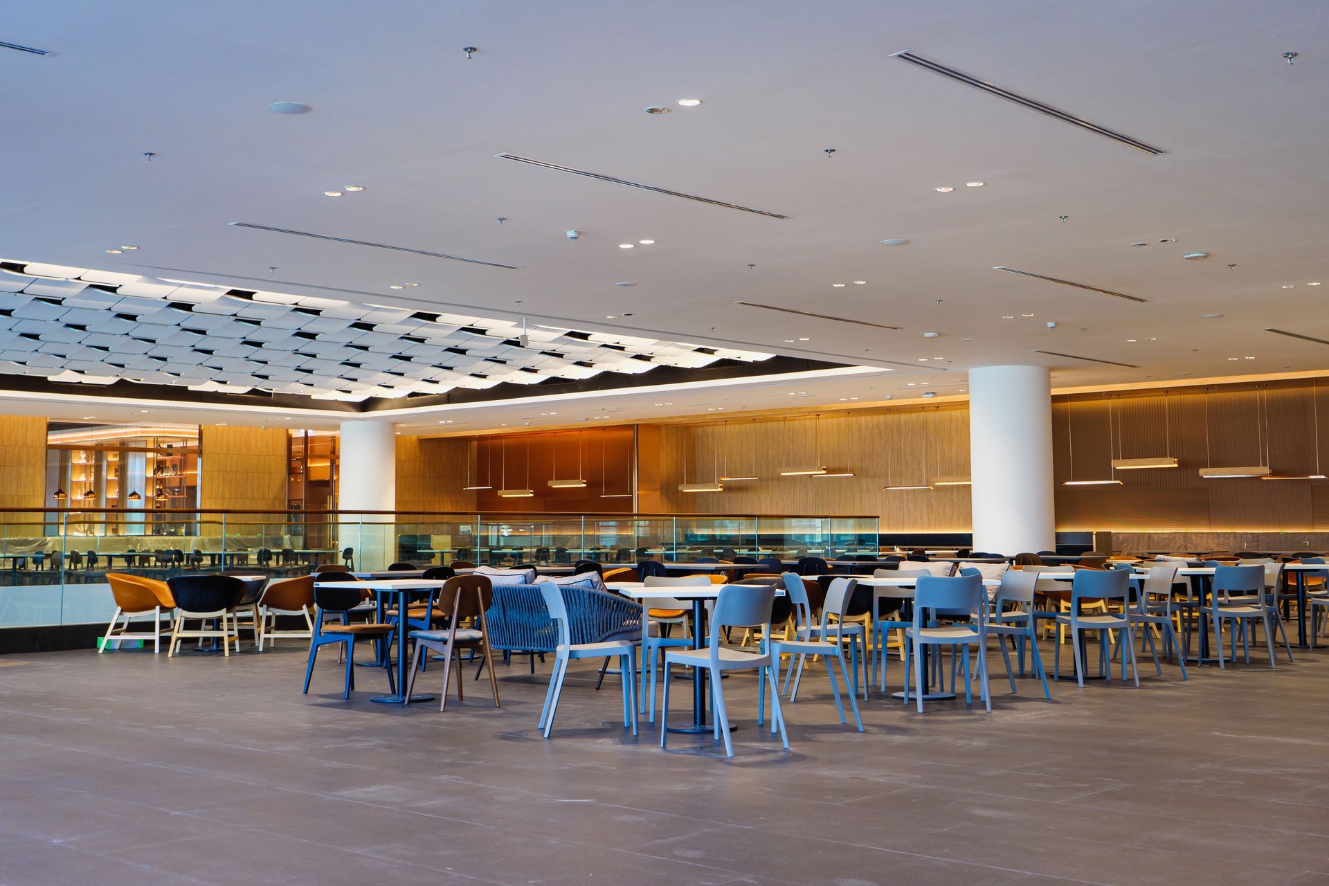 A dining area in a modern building with gray floors, various tables and chairs, white pillars, and a backlit ceiling.
