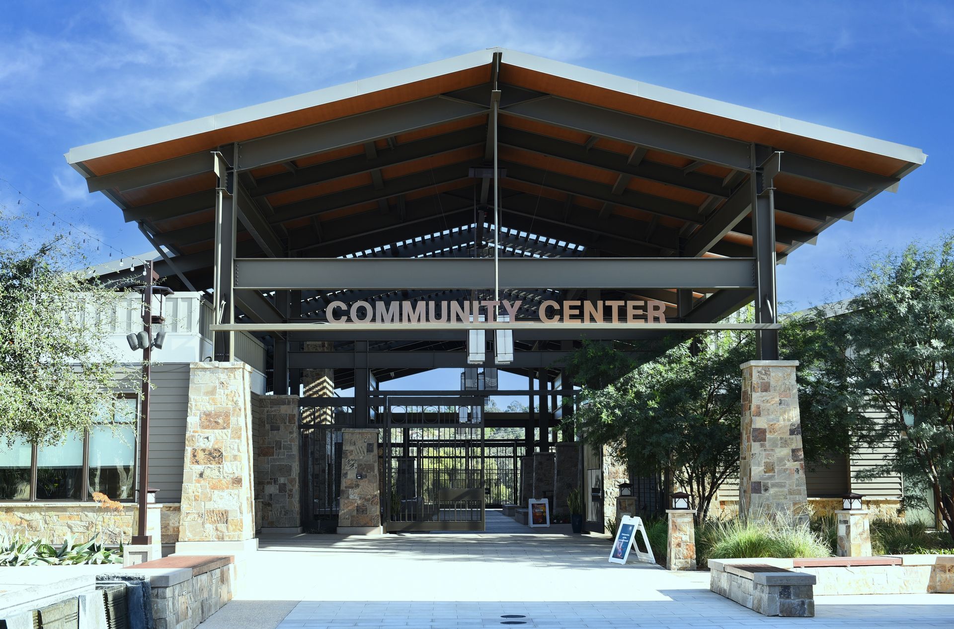The entrance to a modern community center with a high, slanted metal roof, stone pillars, and glass doors under a blue sky.