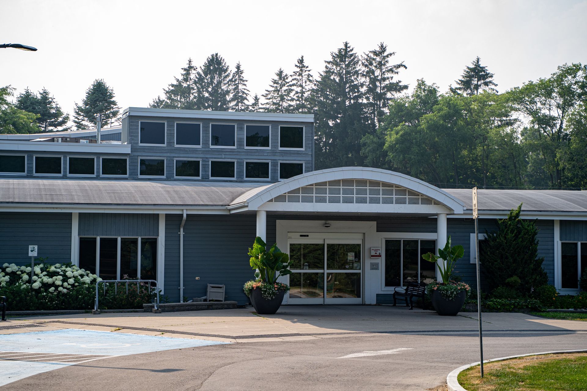 A gray, single-story building with a multi-paneled clerestory roof and a white, arched entryway shaded by trees.