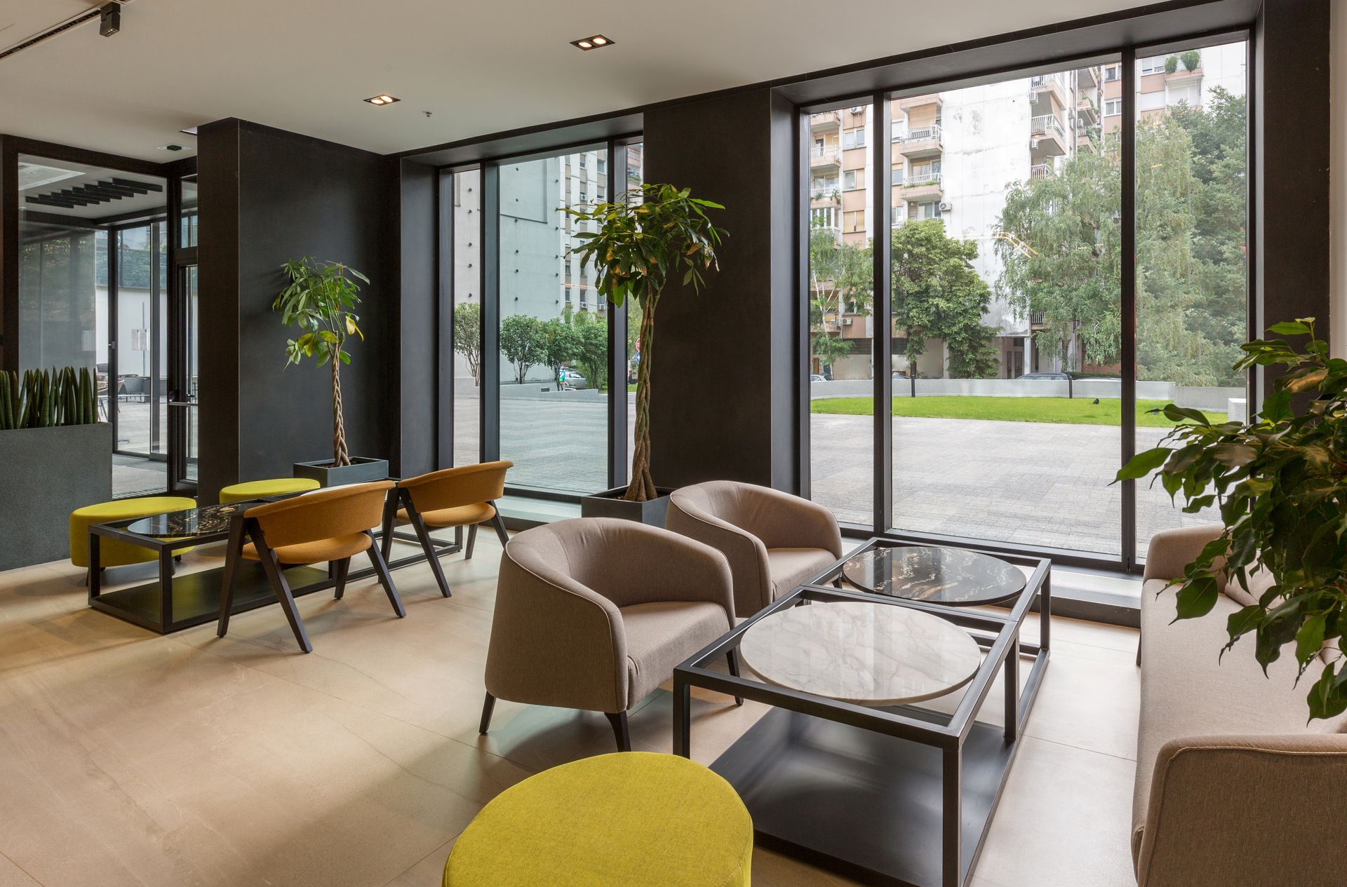 Modern waiting area with beige armchairs, yellow stools, round coffee tables, and potted plants by floor-to-ceiling windows.