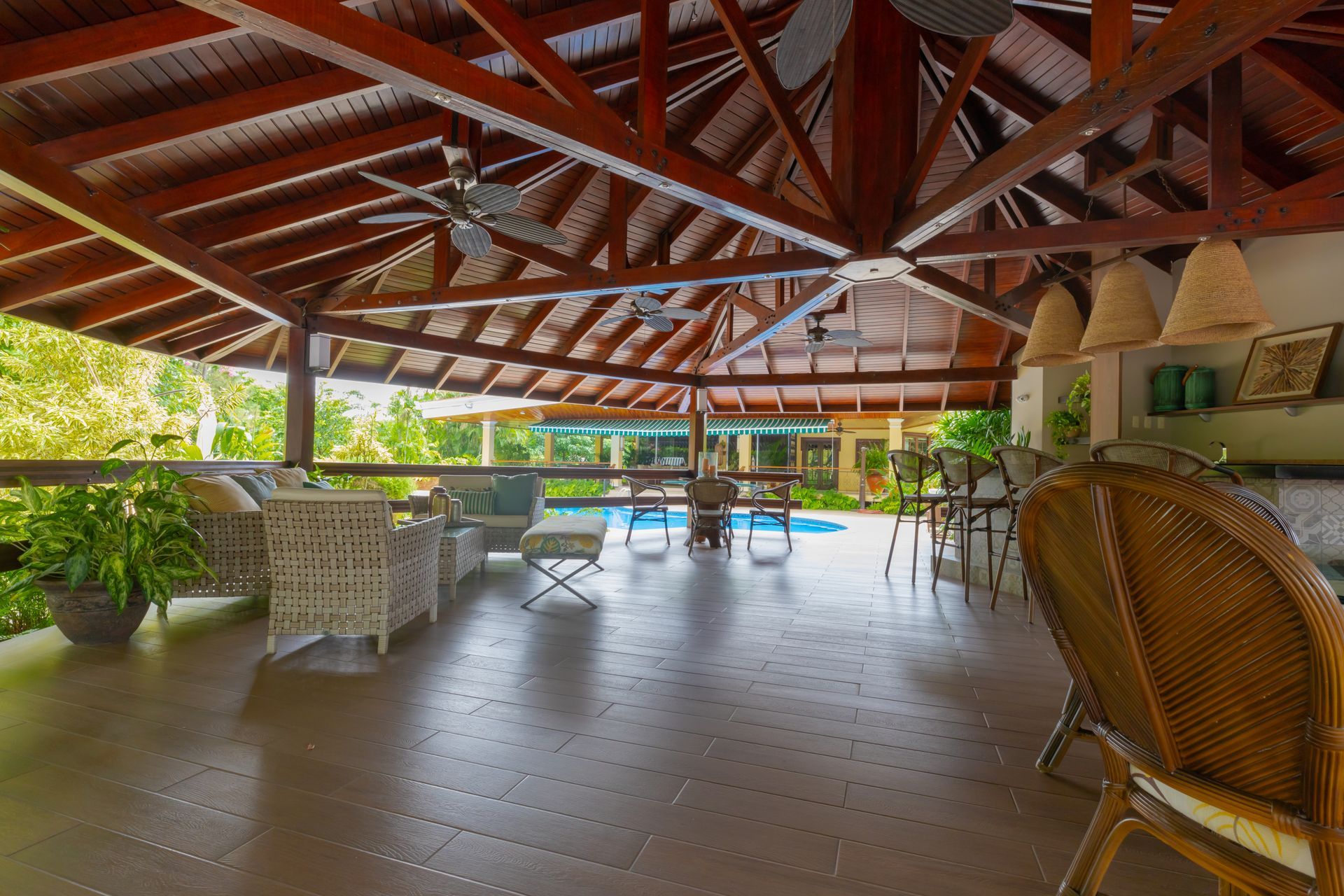 A spacious, covered outdoor patio featuring a vaulted wooden ceiling, wicker furniture, and a view of a swimming pool.