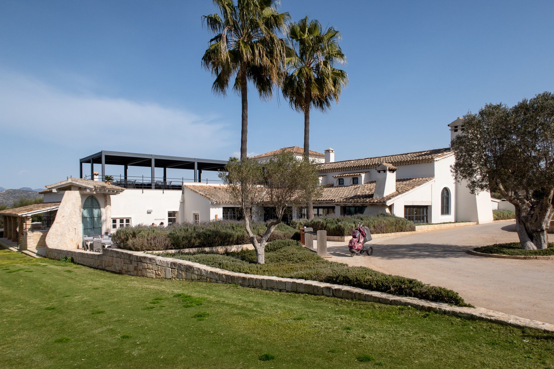 A white building with a patio and palm trees, set behind a stone wall and green lawn under a clear blue sky.