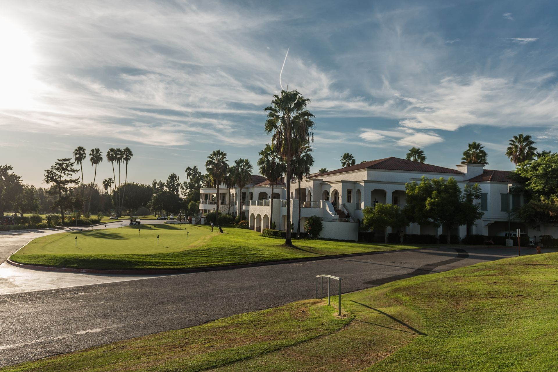 A white Mediterranean-style clubhouse with palm trees sits behind a grassy golf green under a sunny, blue sky.