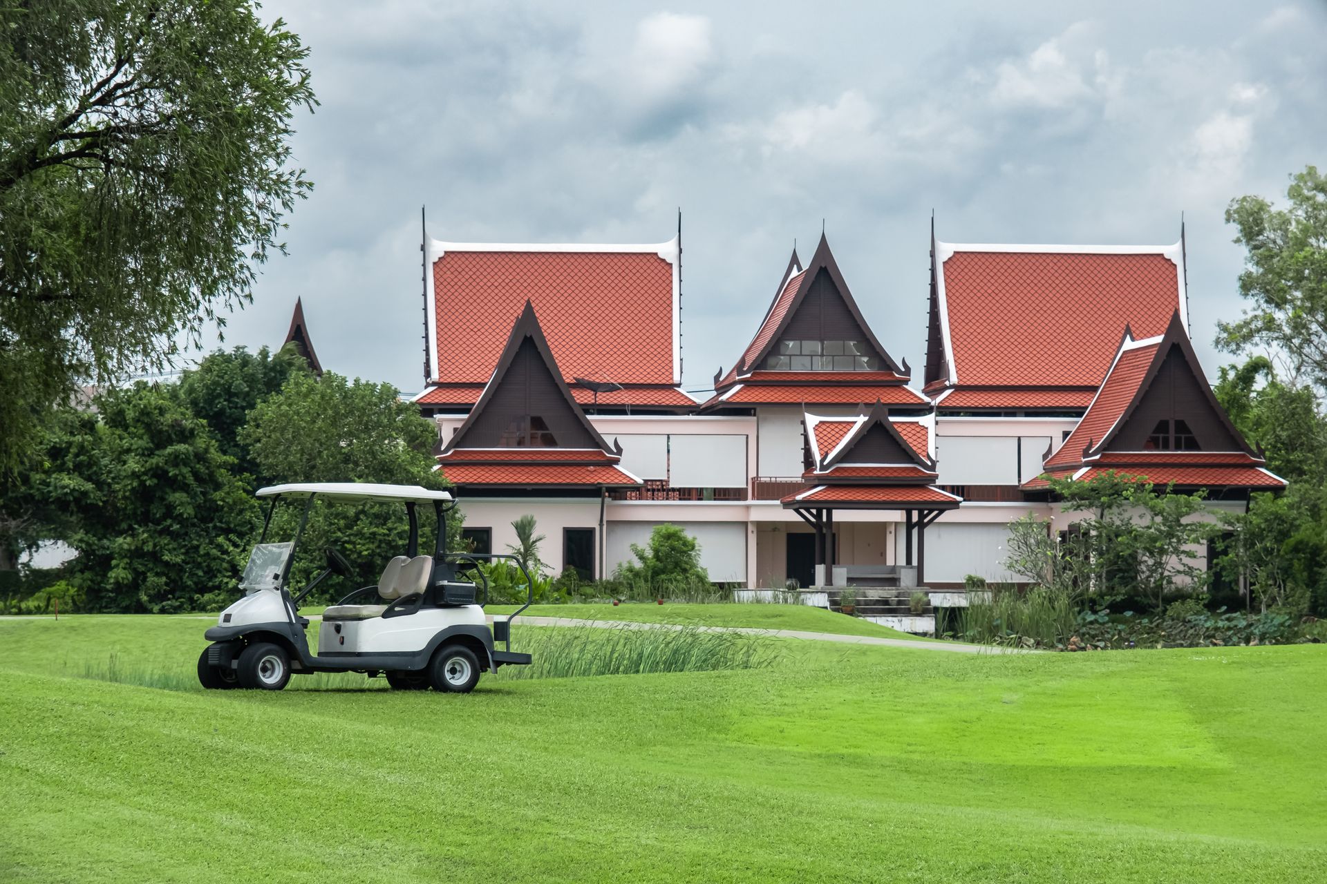 A white golf cart sits on a lush green lawn in front of a traditional Thai-style building with ornate, tiered red roofs.