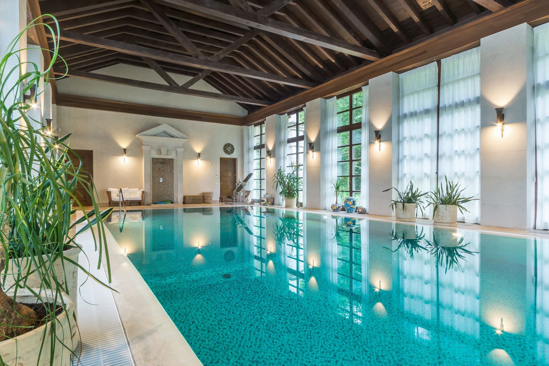 Indoor swimming pool in a room with a vaulted wood-beam ceiling, tall windows, white walls, and potted plants.