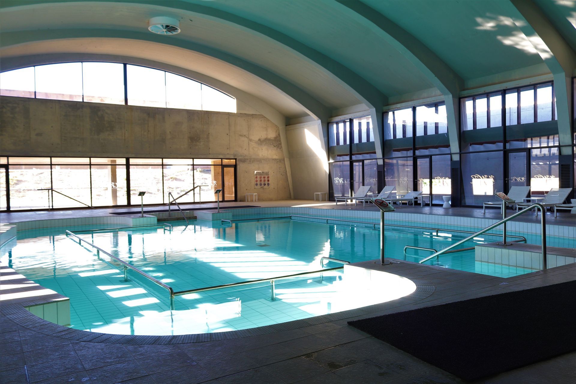 An indoor swimming pool with bright blue water, large windows, and a curved ceiling.