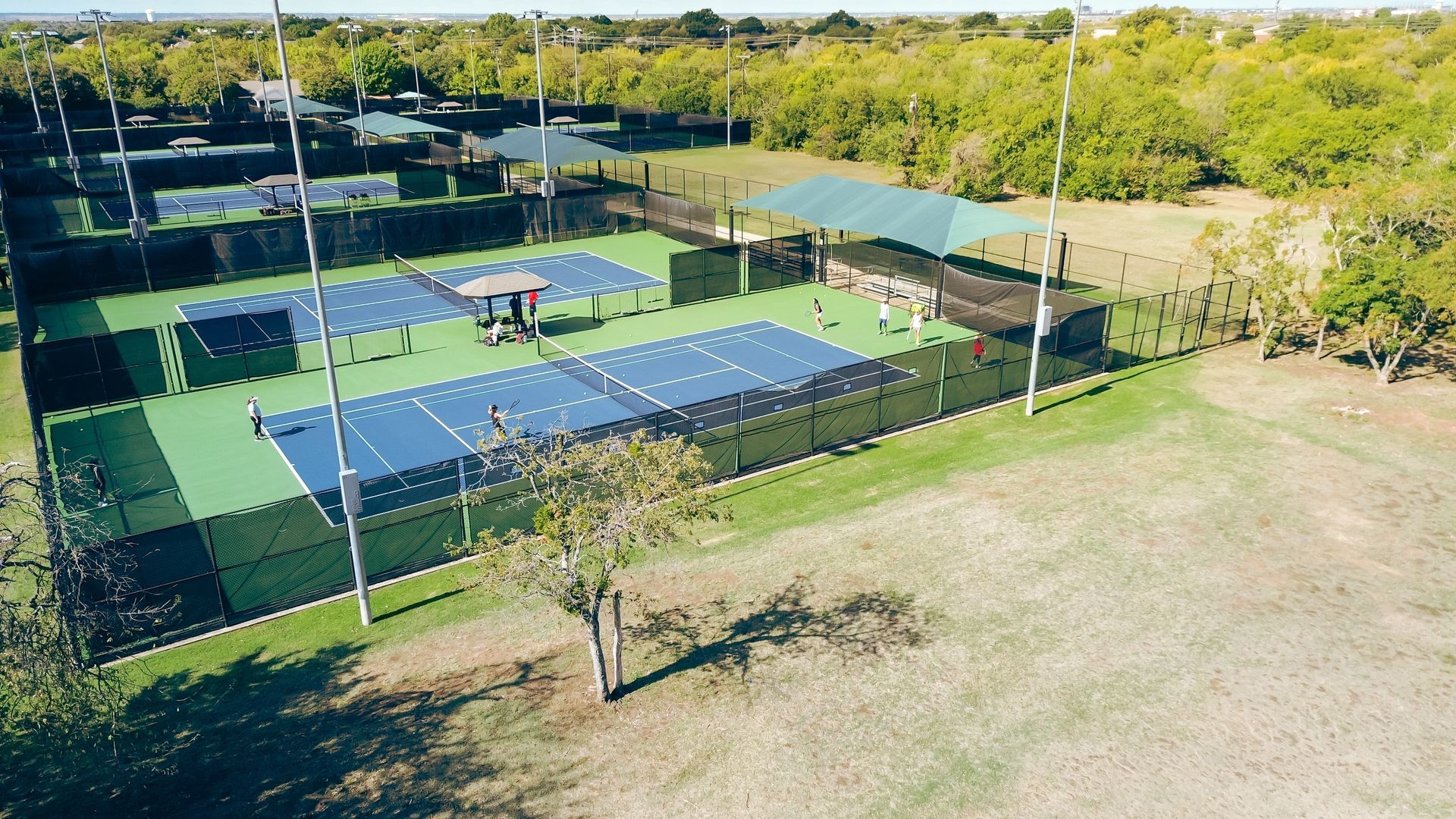 An elevated view of multiple blue pickleball courts enclosed by black fencing, adjacent to green trees and open land.