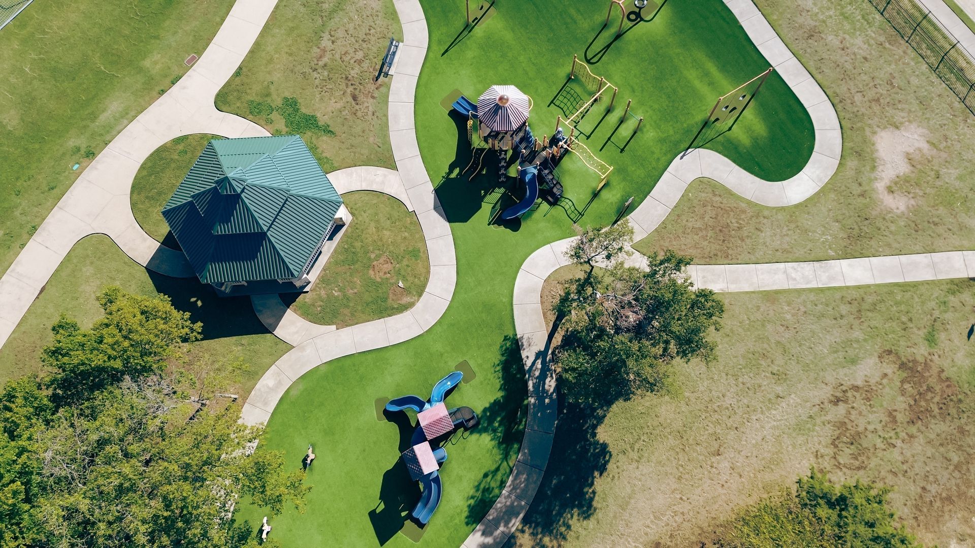 An aerial view of a park with a green-roofed gazebo, two playgrounds, and winding concrete paths on a sunny day.