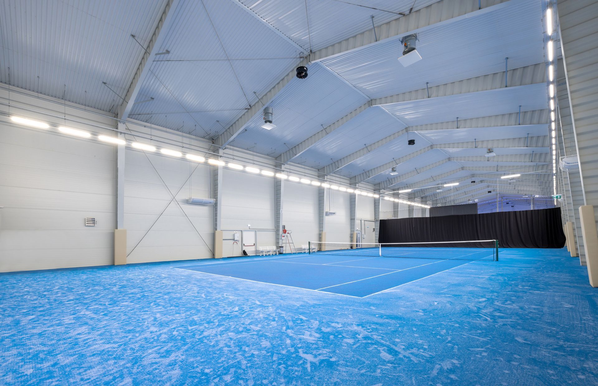 An indoor sports facility with bright blue flooring and a tennis or padel court marked with white lines.