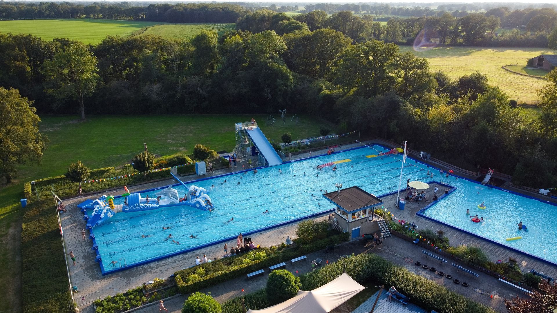 An aerial view of an outdoor swimming pool complex with a slide, an inflatable obstacle course, and grassy surroundings.
