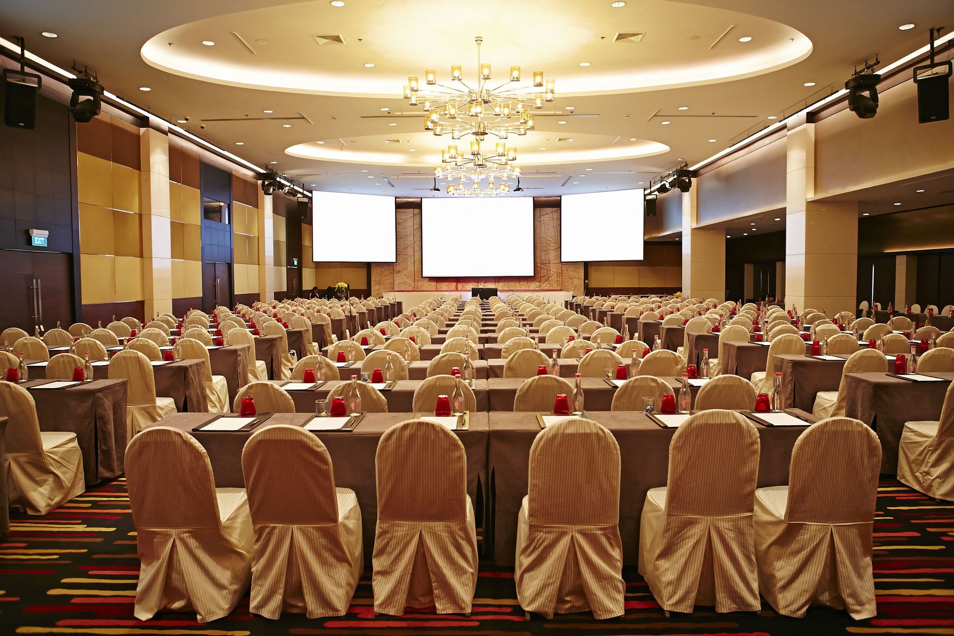 A large, empty conference room with rows of tables, beige chairs, and three large screens on the front wall.