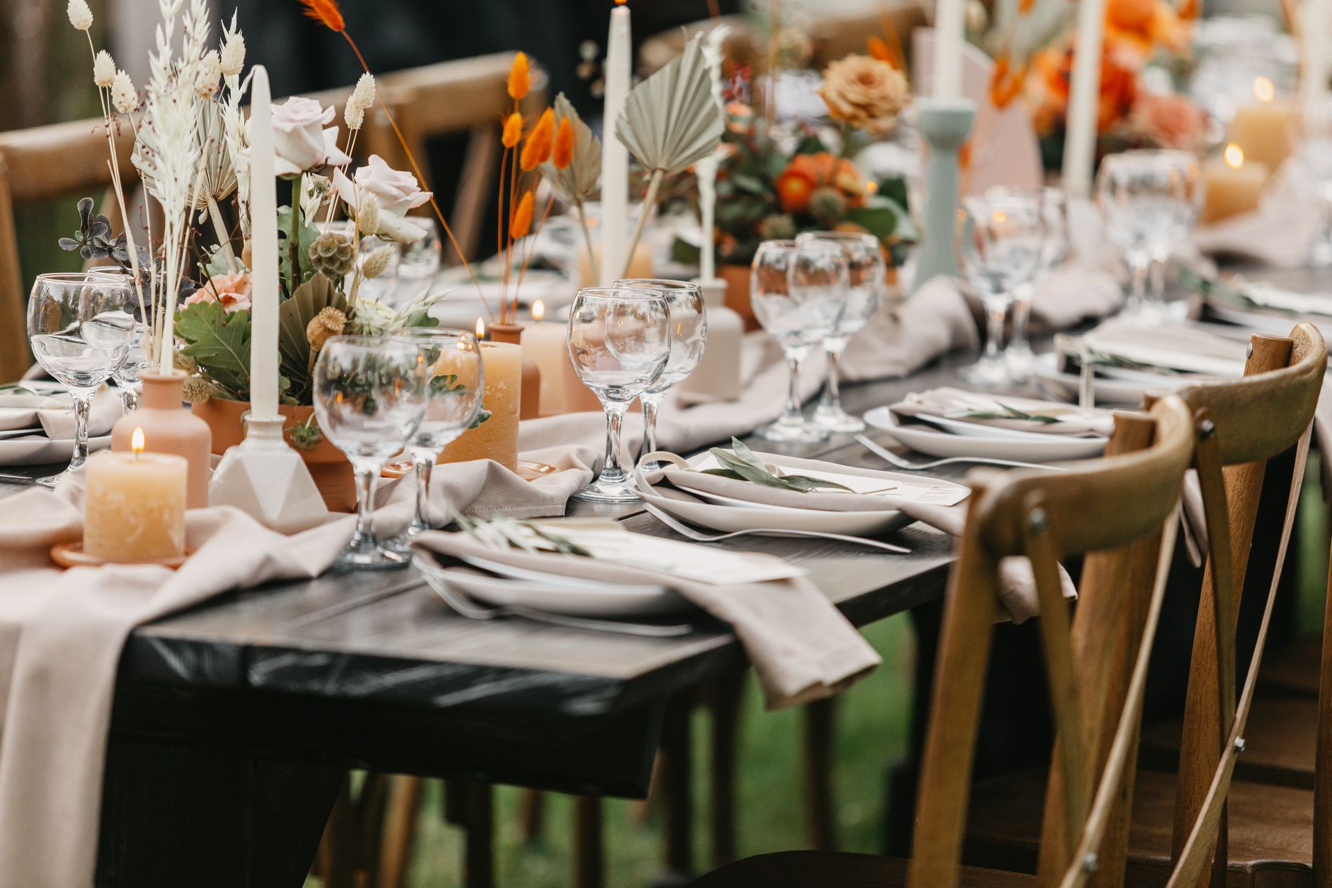 A long outdoor dining table set for an event with beige runners, pillar candles, floral centerpieces, and wooden chairs.