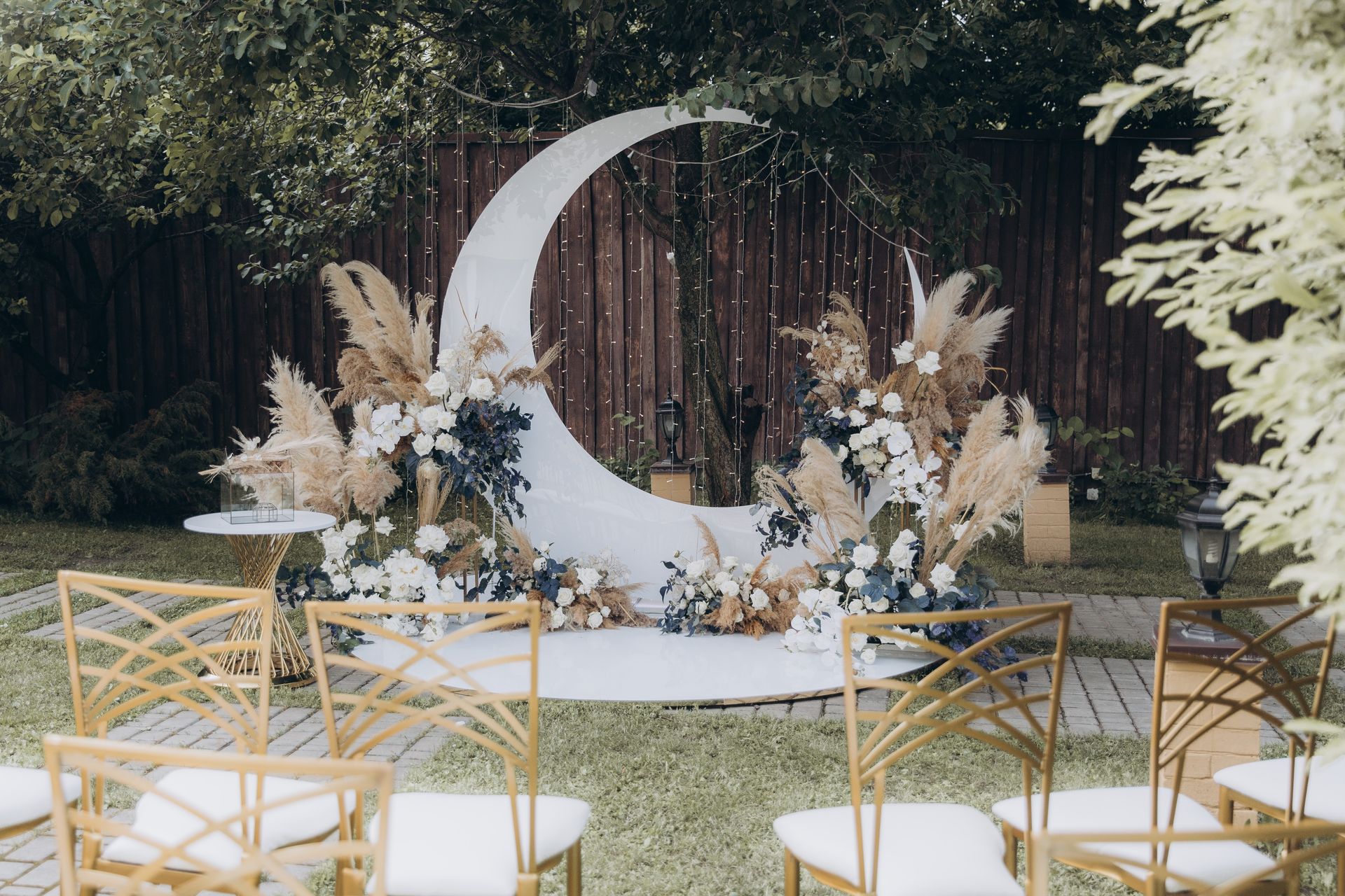 A crescent moon wedding backdrop decorated with pampas grass and white flowers, with gold chairs arranged in a garden.