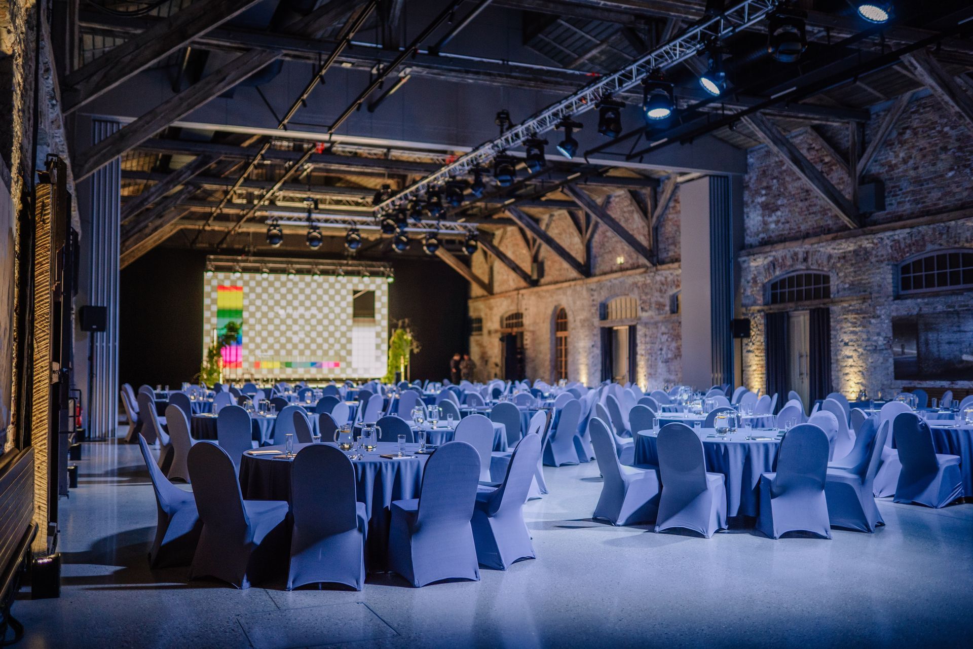 A spacious event hall with rustic stone walls, wooden rafters, and many round tables set for a formal gathering.
