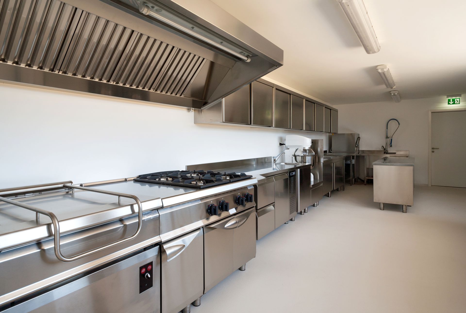 A modern, clean, stainless steel commercial kitchen with a gas stove, range hood, overhead cabinets, and prep tables.