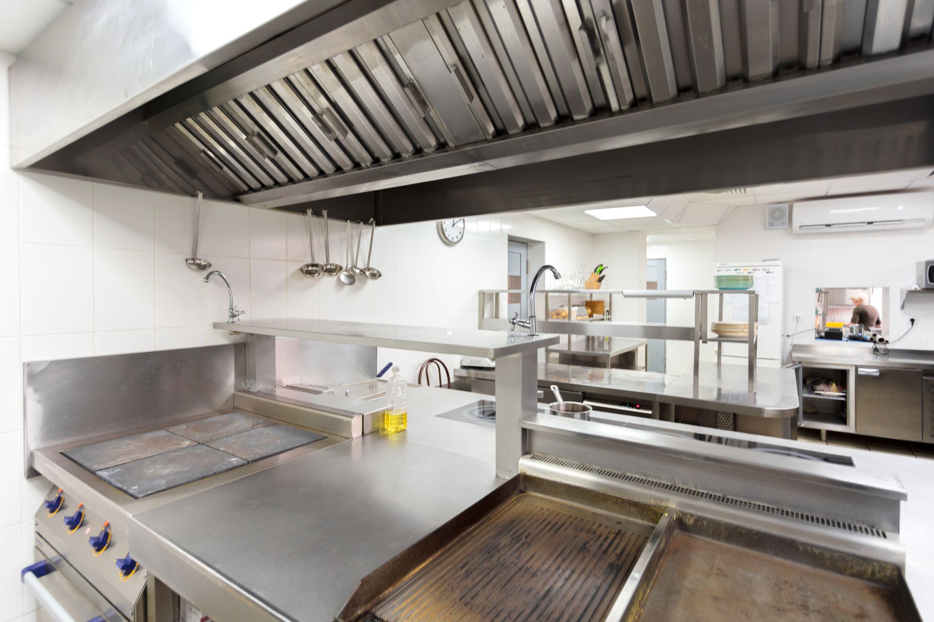 A professional stainless steel commercial kitchen with griddles, work surfaces, and a large overhead exhaust hood.
