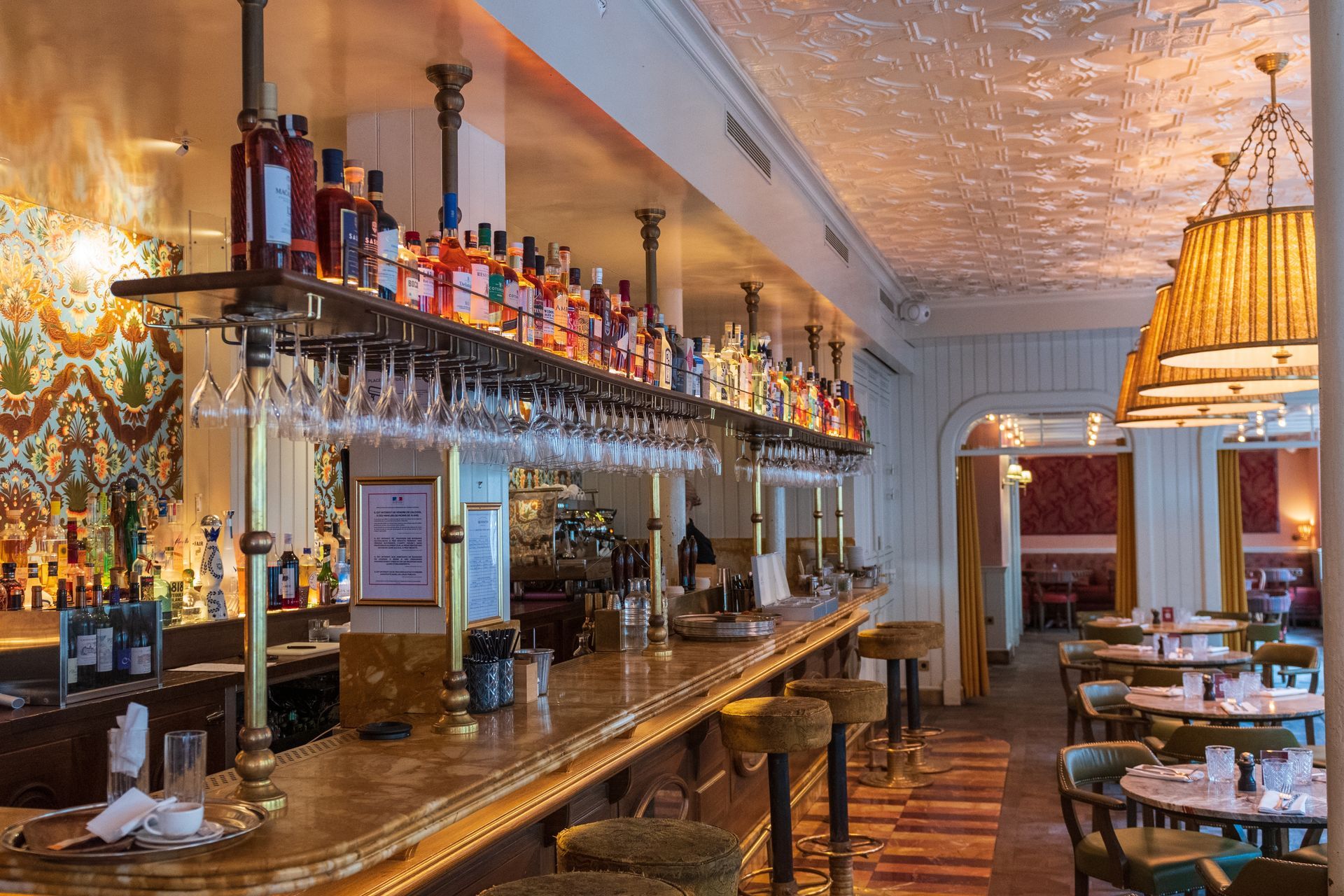 An elegant, well-lit restaurant bar featuring a long gold counter, hanging wine glasses, and rows of bottles on shelves.