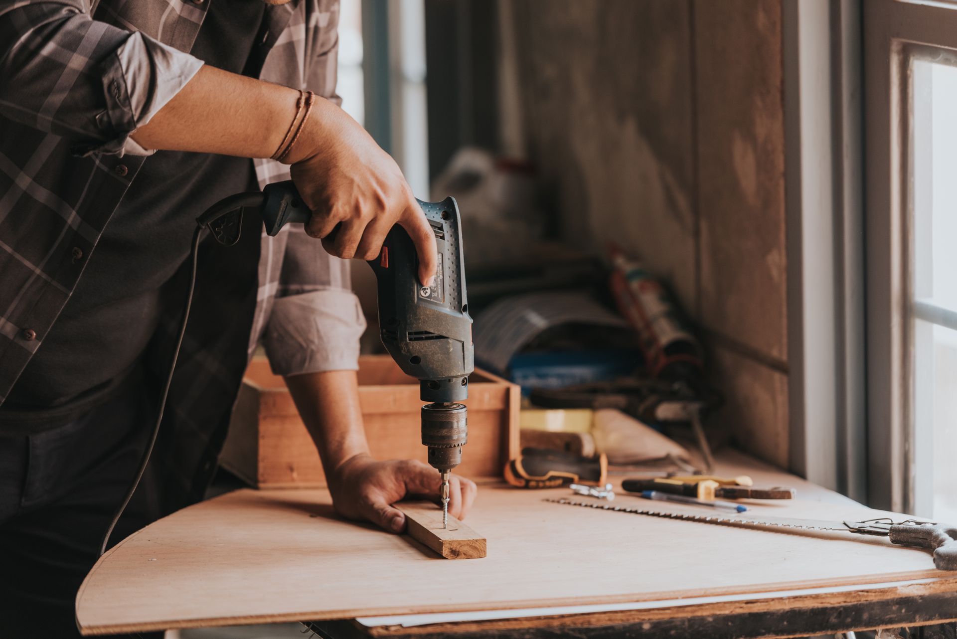 A person uses a power drill on a small wooden block at a workbench in a workshop.