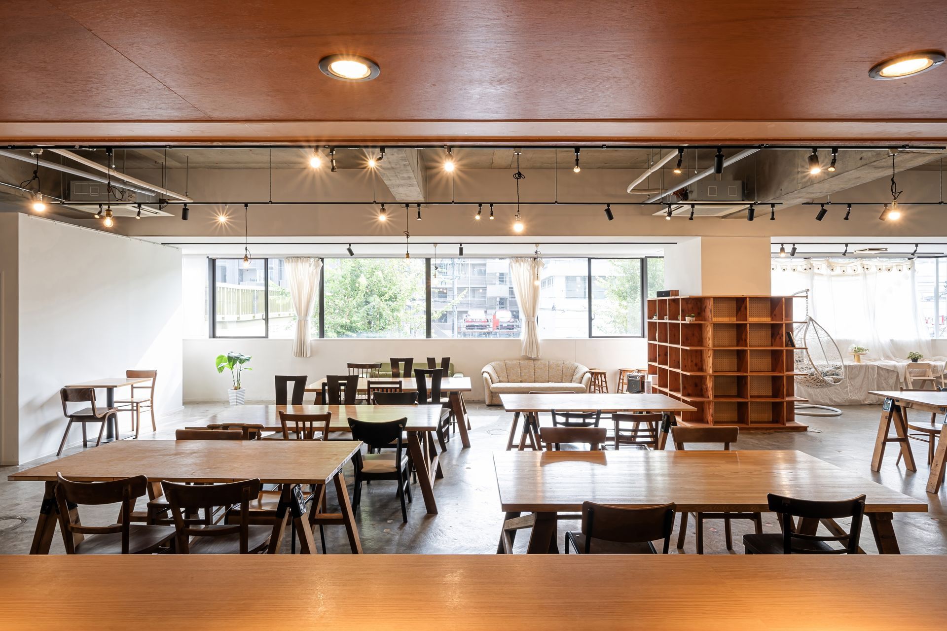 A bright, open-plan workspace or cafe with wooden tables, chairs, and large windows, viewed from a slightly elevated spot.