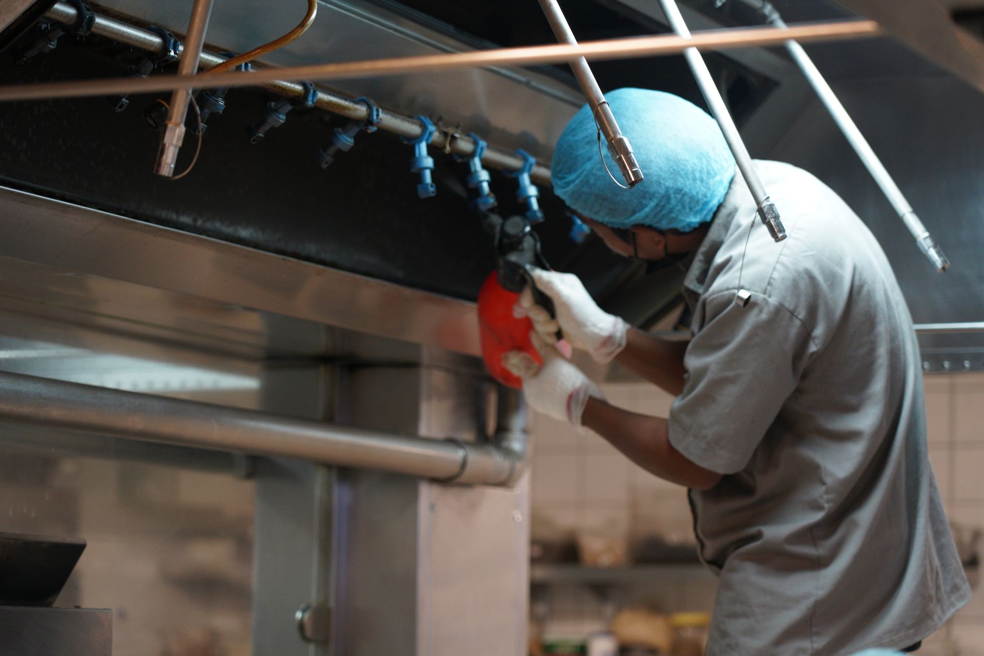 A maintenance worker in a hairnet and gloves cleans the ventilation nozzles inside a commercial kitchen hood.