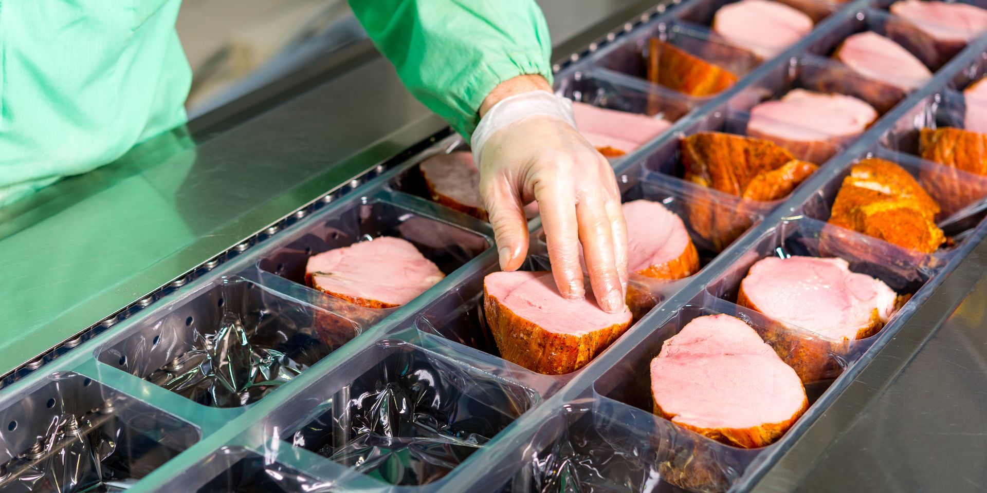 A gloved hand places portions of seasoned meat into individual plastic trays on a food processing conveyor belt.