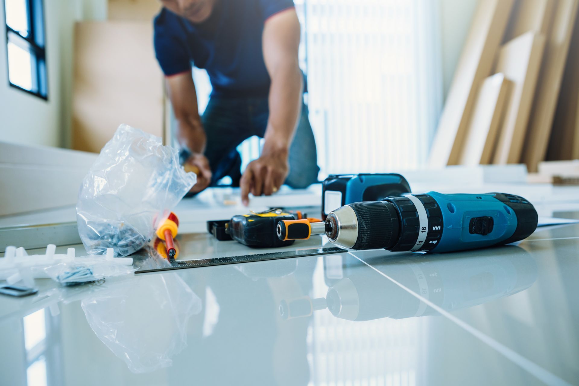 A person works on a home project on a bright floor, with a blue cordless drill and tools laid out in the foreground.