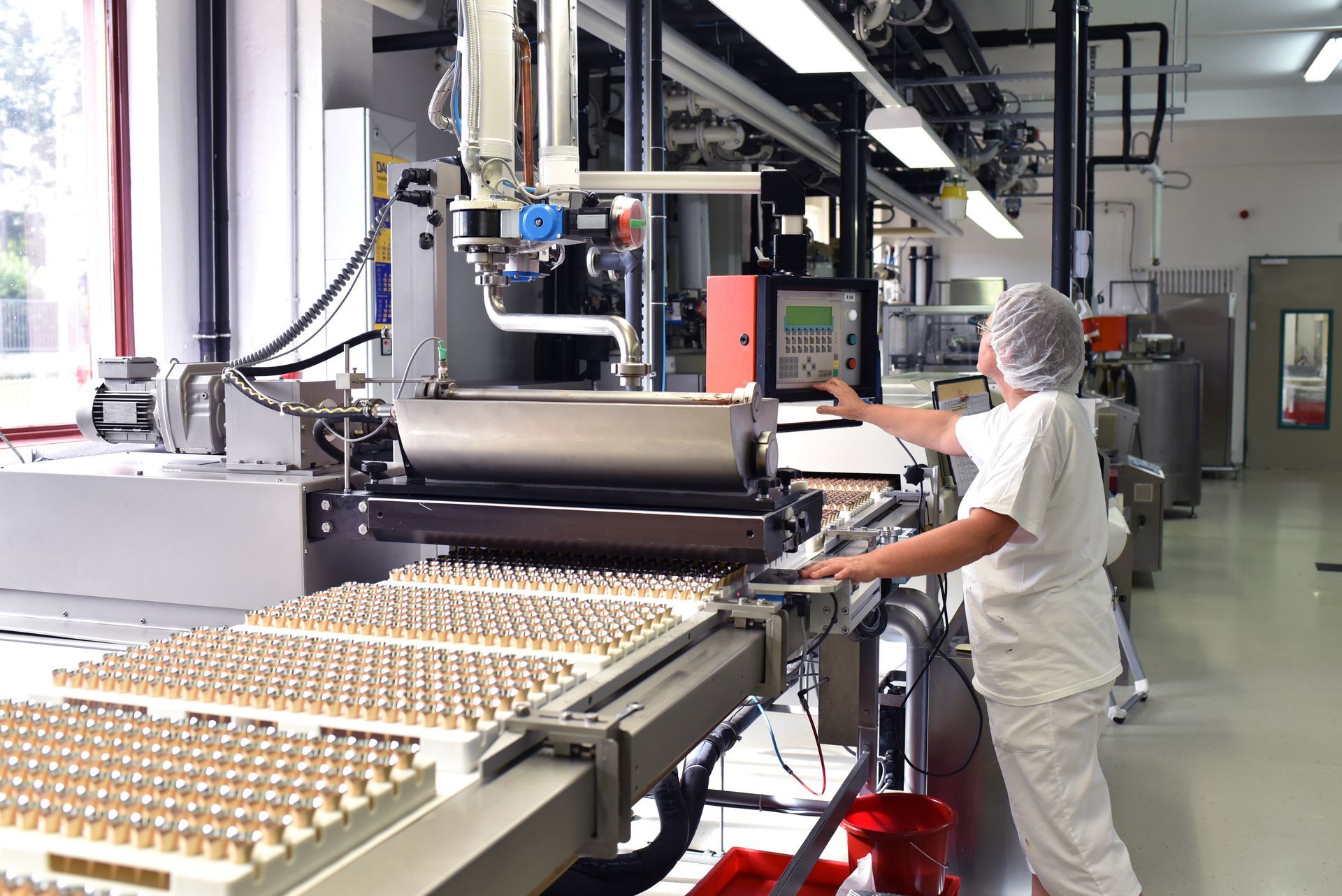 A worker in white uniform and hairnet operates an industrial packaging machine filled with small glass vials.
