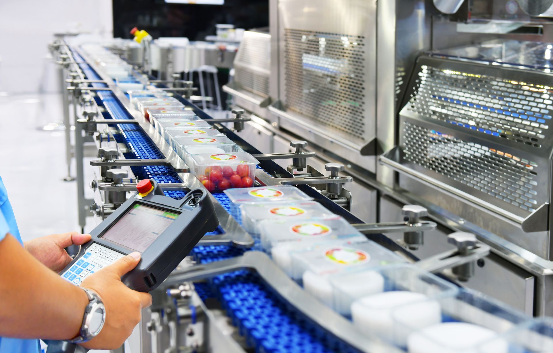 A person holding a handheld control device monitoring a blue conveyor belt carrying food containers in a facility.