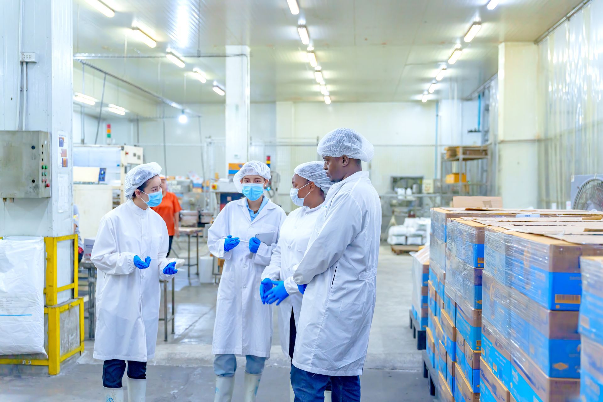 Four food factory workers in white lab coats, masks, and blue gloves stand in a warehouse discussing products.