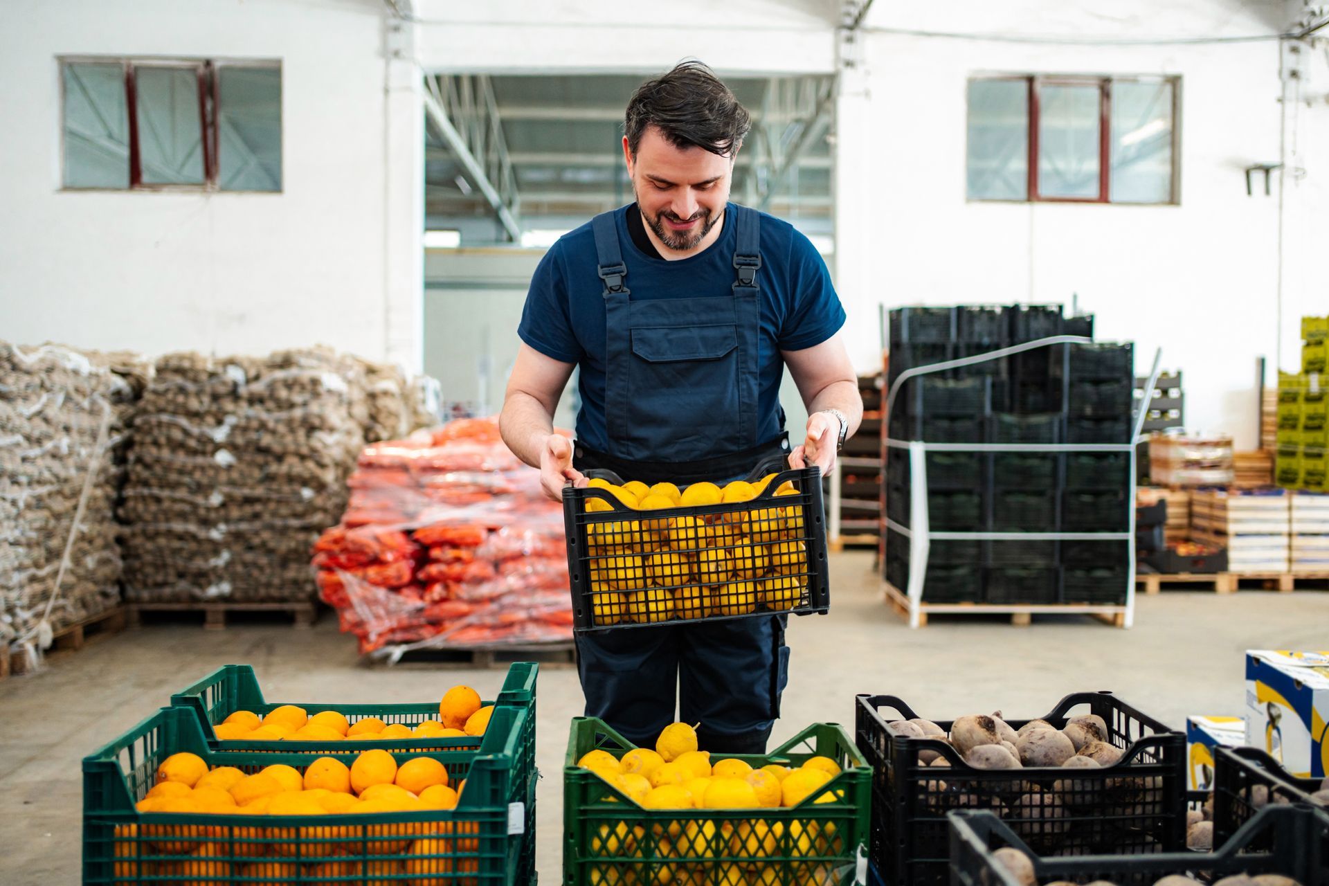 A worker in blue overalls smiles while holding a crate of yellow citrus fruit in a warehouse filled with produce crates.
