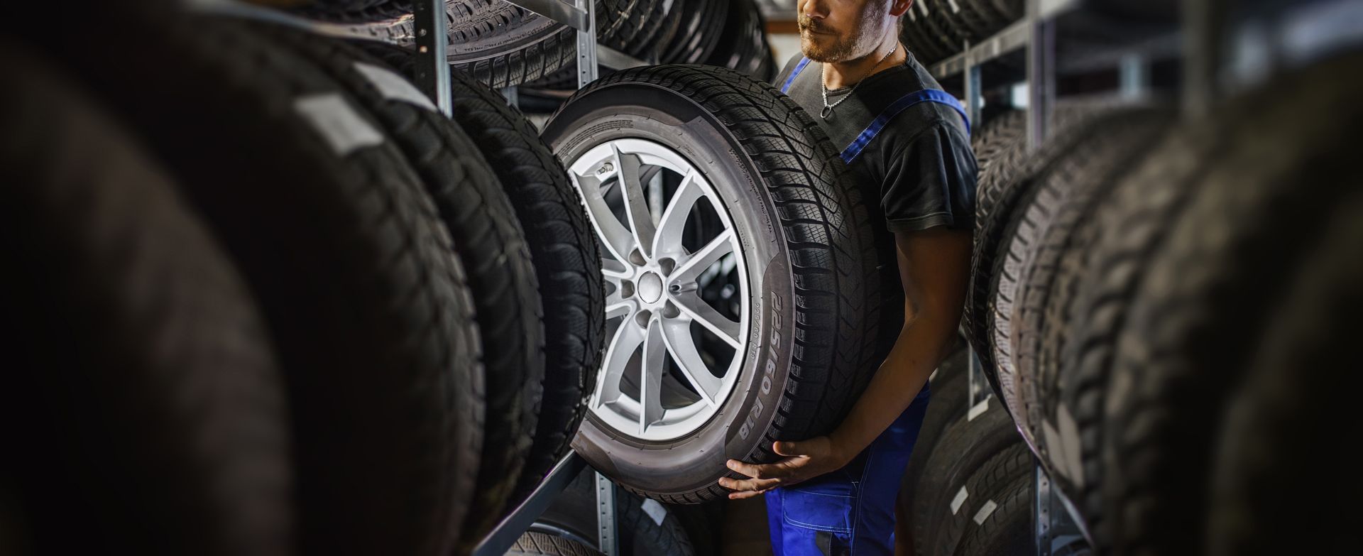 A person in work clothes lifting a tire on a rim from a storage rack in a workshop.