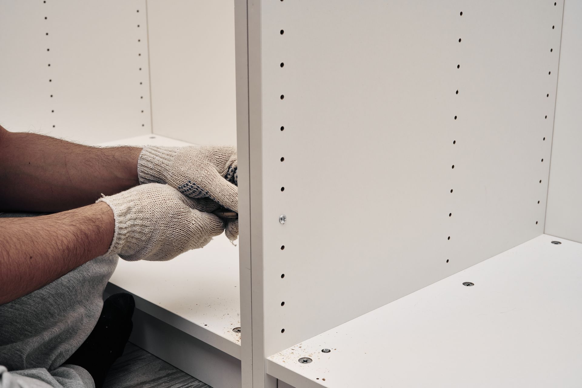 A person wearing knit work gloves assembles white furniture panels by inserting screws into pre-drilled holes.
