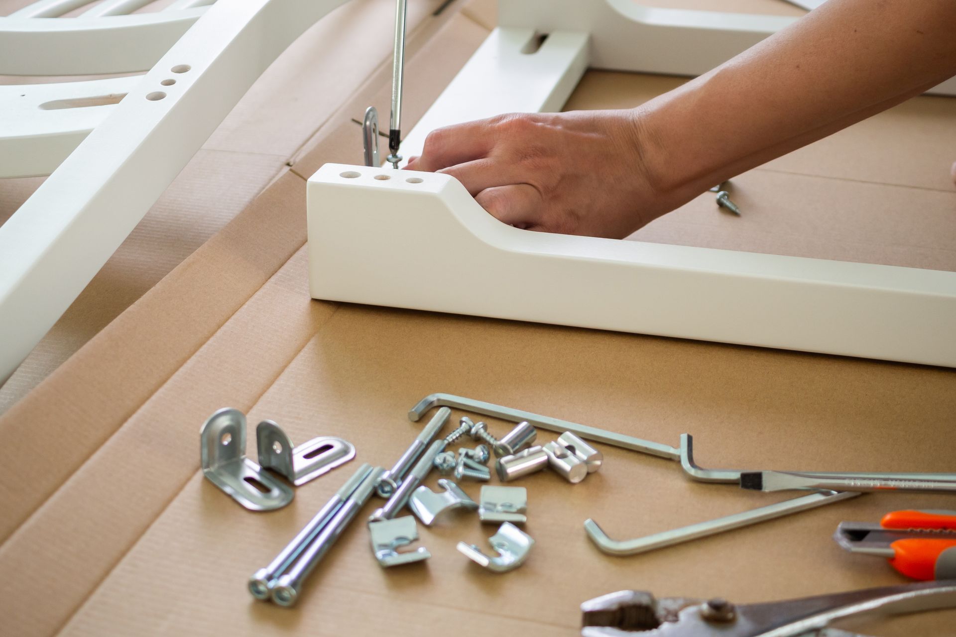 A person assembles white wooden furniture on a cardboard surface with various metal hardware and tools scattered nearby.