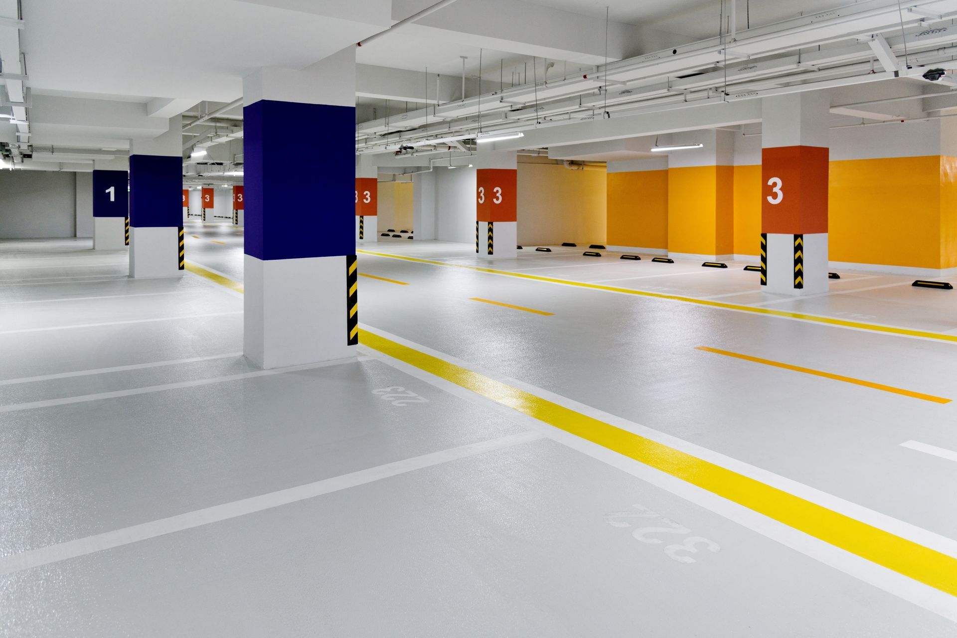 Empty underground parking garage with white pillars, blue and orange accents, and yellow lane markings on gray floors.