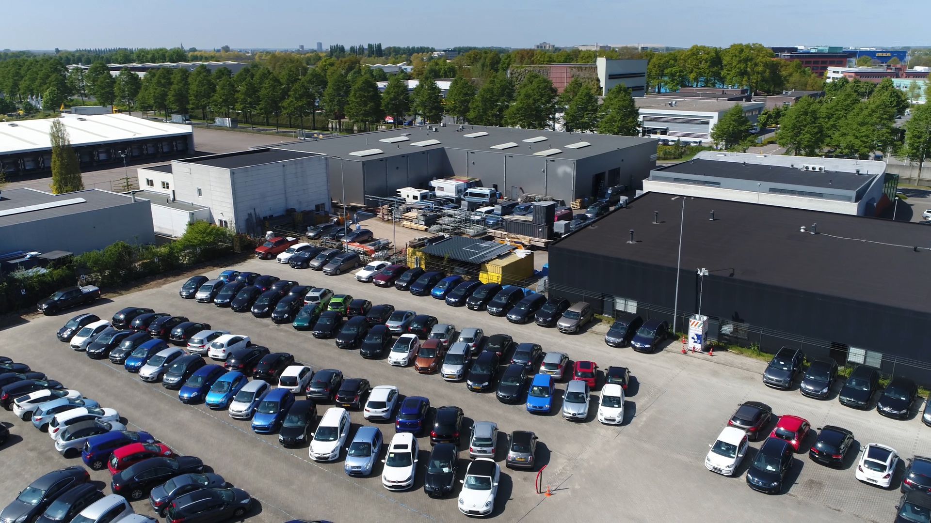 An aerial view of a large parking lot filled with many cars parked in rows next to industrial buildings on a sunny day.
