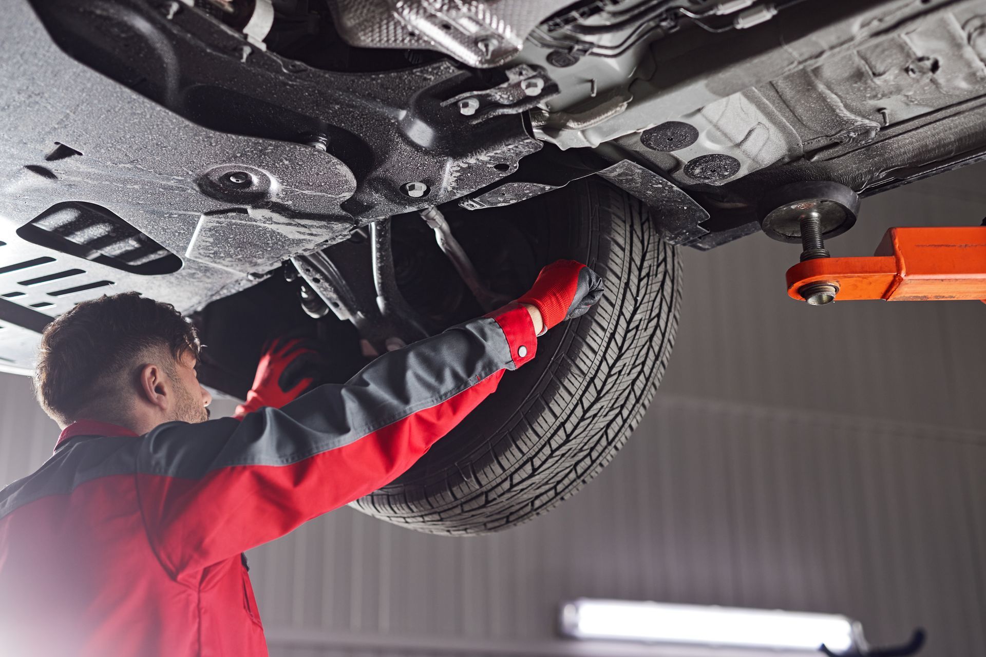 A mechanic in a red and gray uniform works on the raised tire and suspension of a car in a garage.