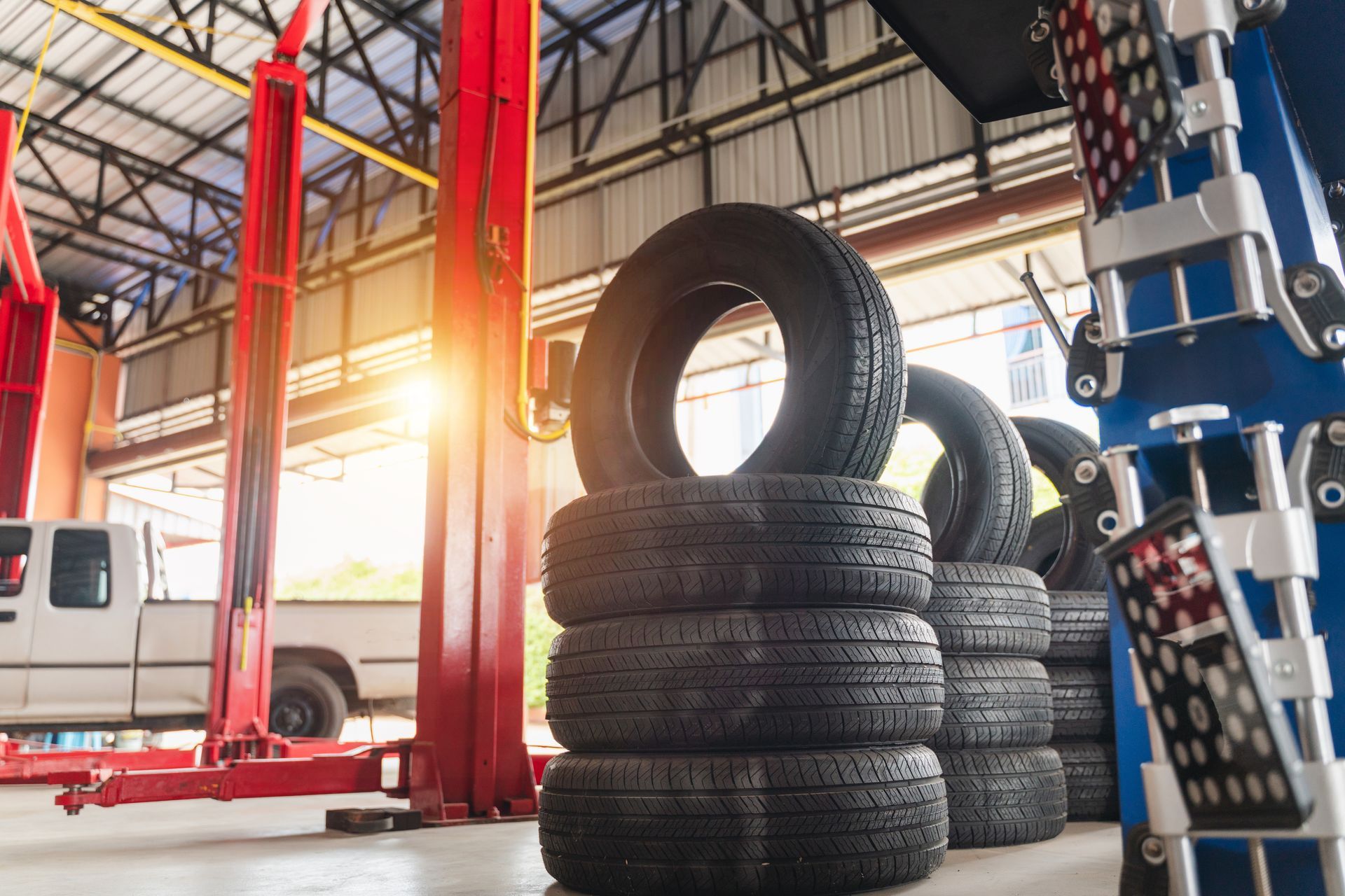A stack of black tires in a brightly lit automotive repair shop with a vehicle lift and alignment equipment in view.