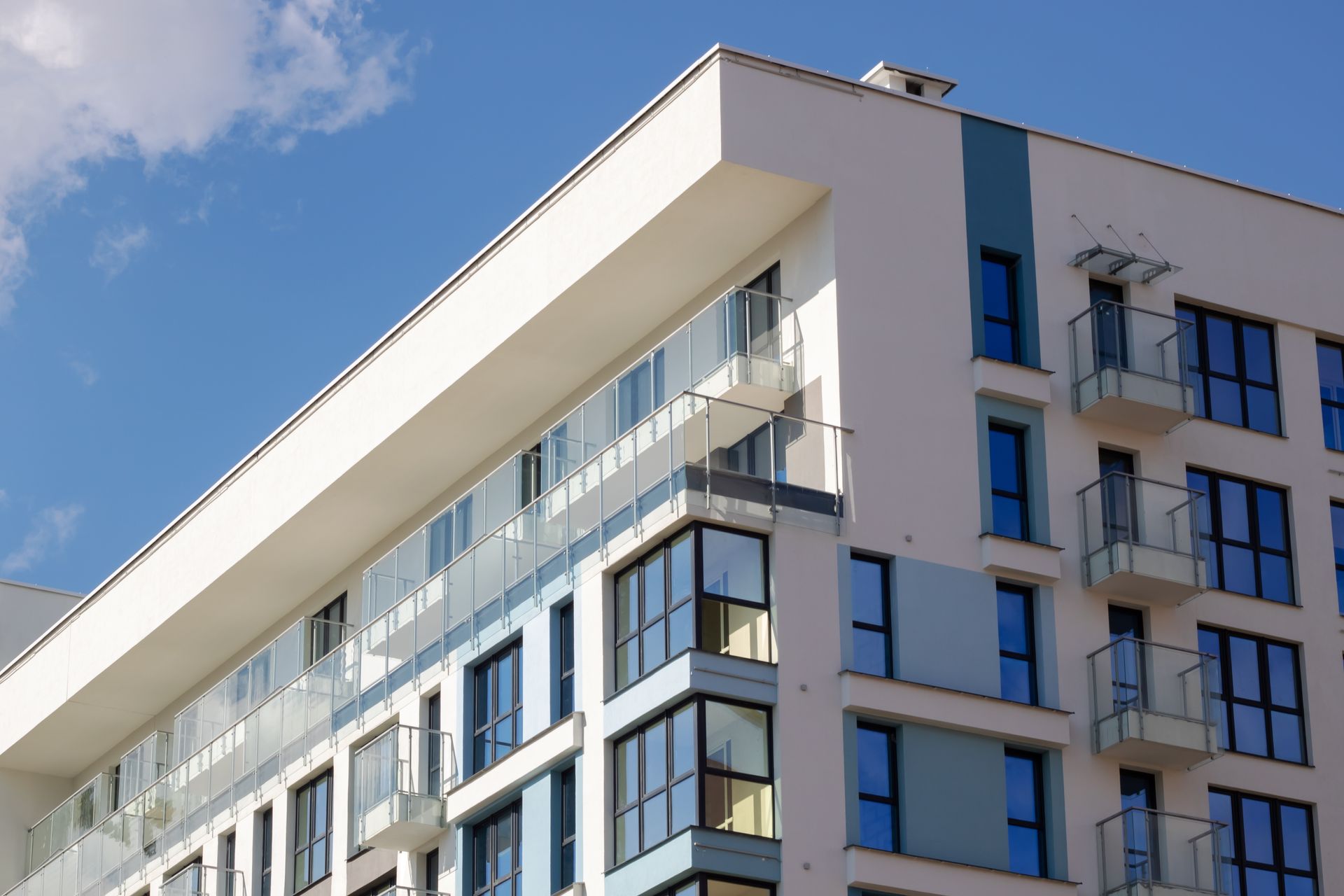 A modern, multi-story apartment building with a white facade, blue accents, glass balconies, and many large windows.