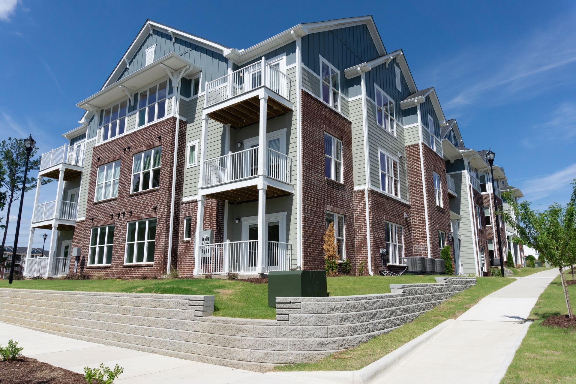 Multi-story apartment building with brick and siding exterior, balconies, and a stone retaining wall on a sunny day.