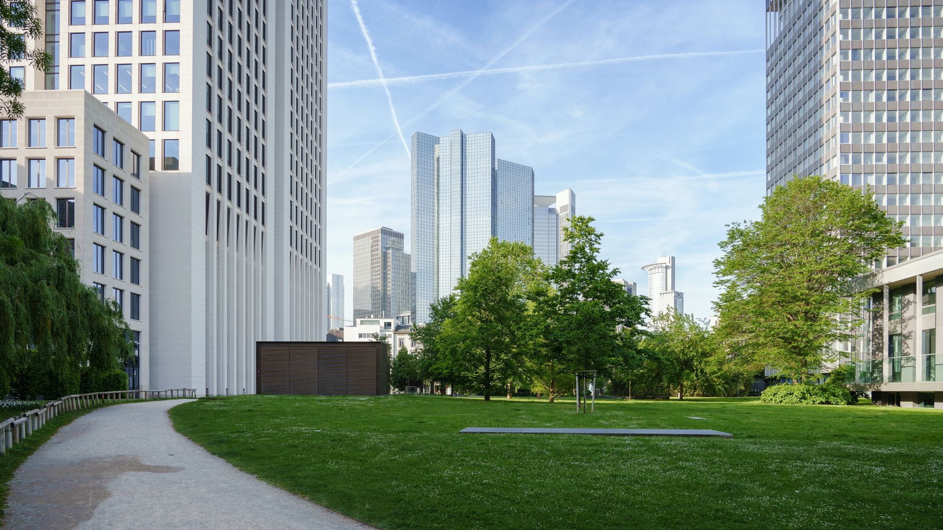 A park path leads toward a dark rectangular structure, framed by modern glass skyscrapers under a blue sky.