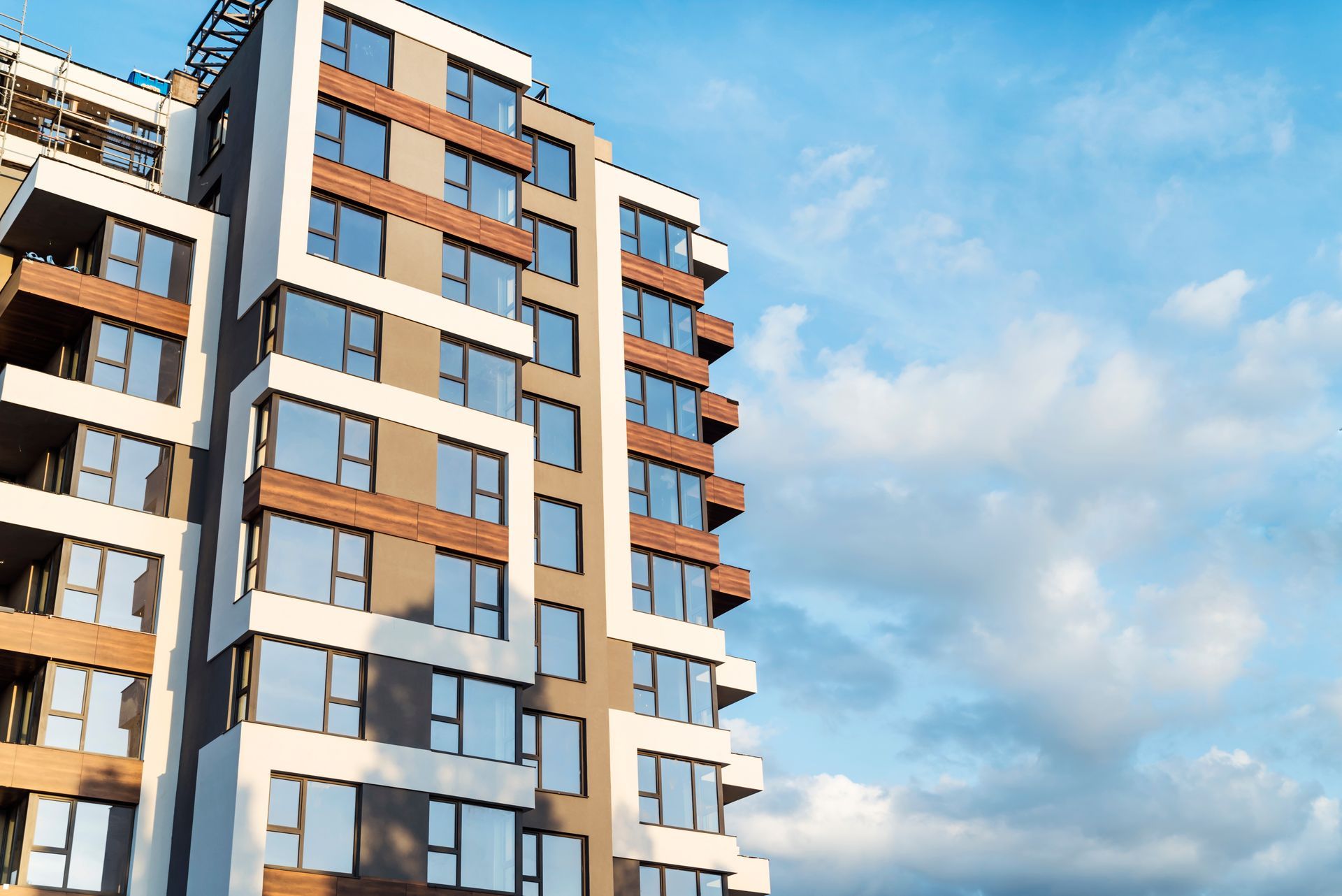A tall, modern apartment building with white frames and brown accents against a blue sky with soft, scattered clouds.