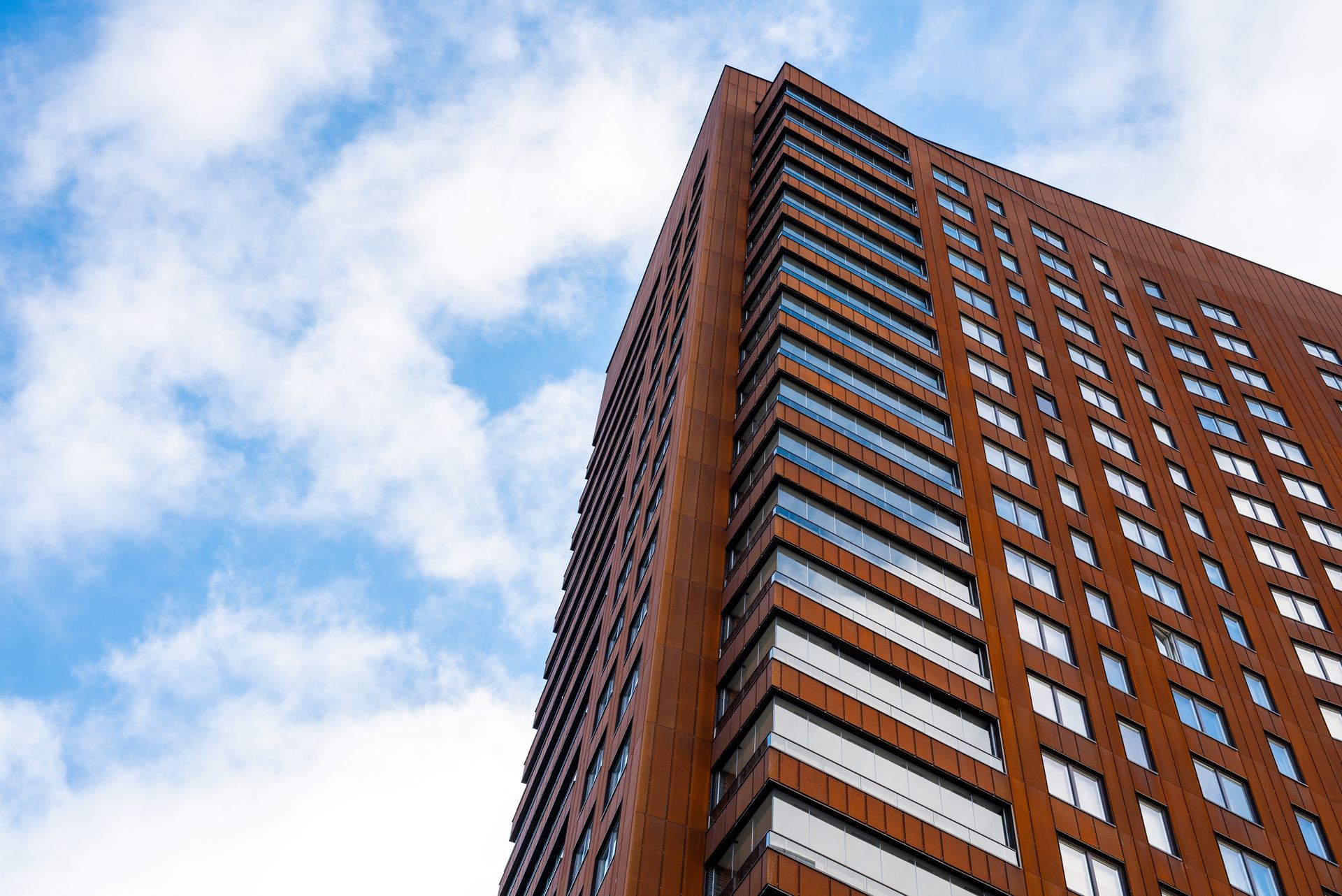 A tall, modern apartment building with a rusted metal facade against a bright blue sky with white clouds.