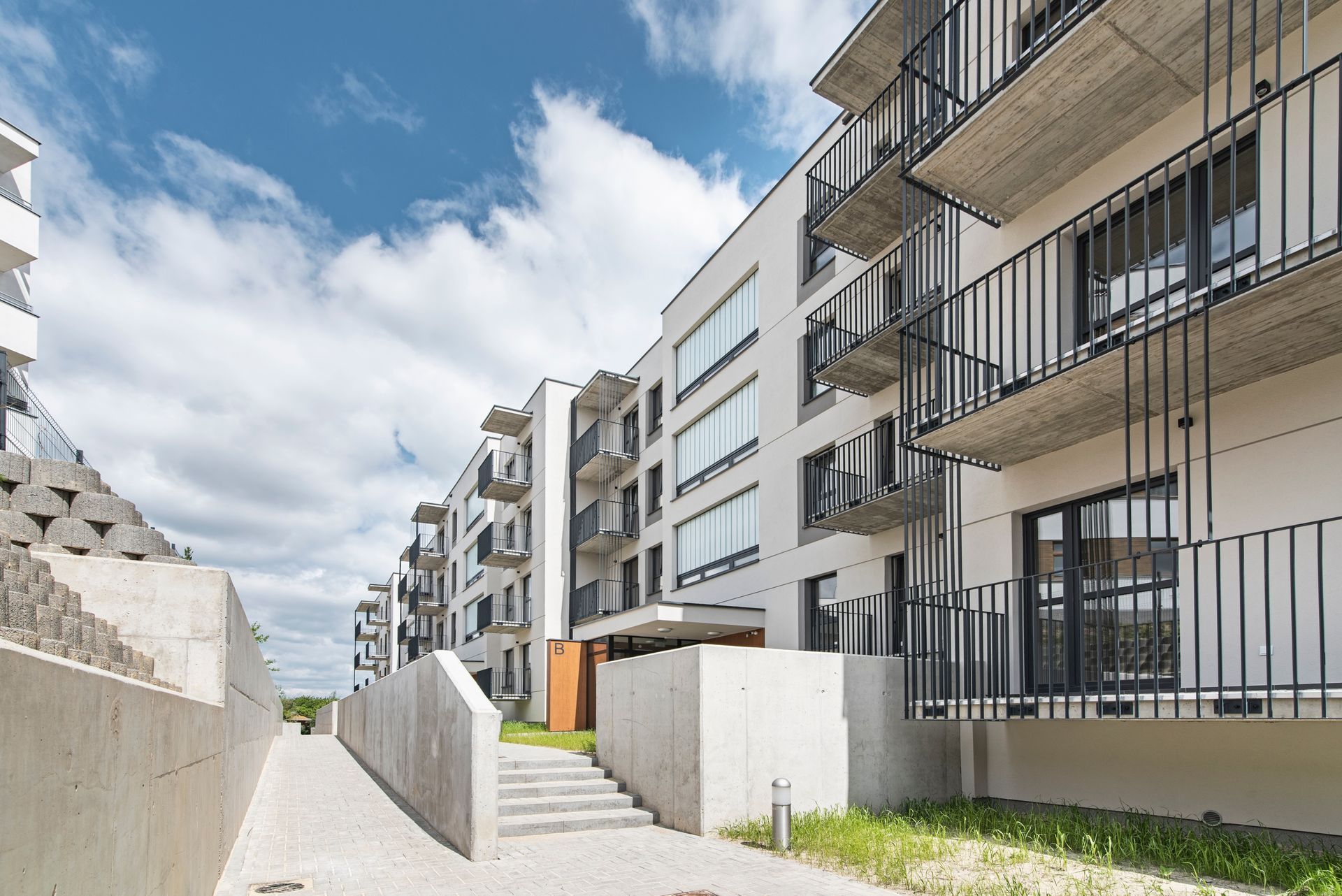 A modern, white apartment complex with stacked balconies and concrete outdoor stairs under a cloudy blue sky.