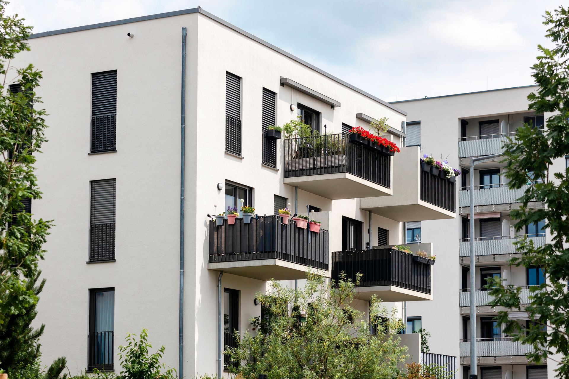 Modern white apartment building with tiered balconies featuring dark railings and potted plants, set against a blue sky.