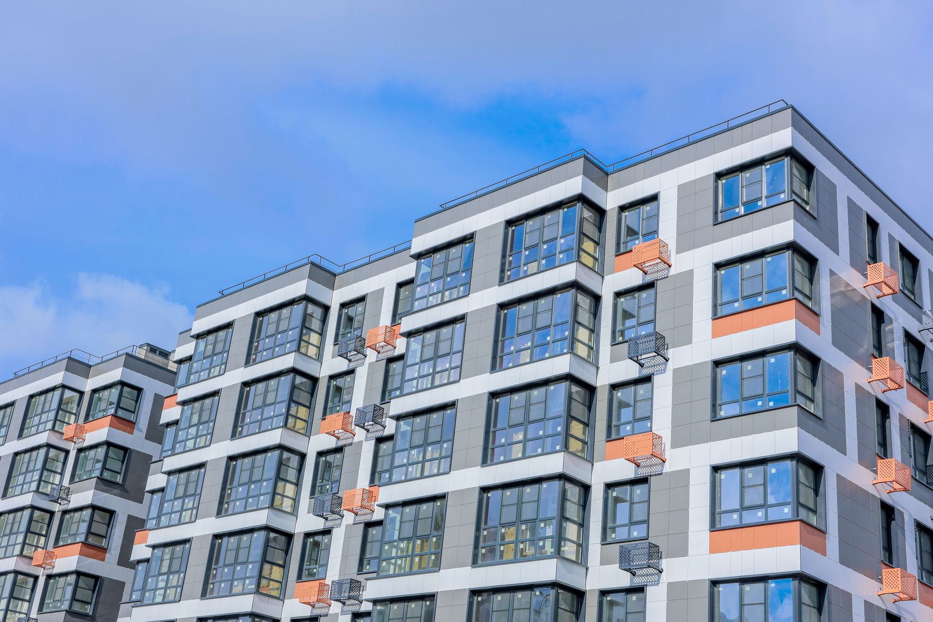 A modern multi-story apartment building with a white and gray facade, large windows, and small orange balcony accents.