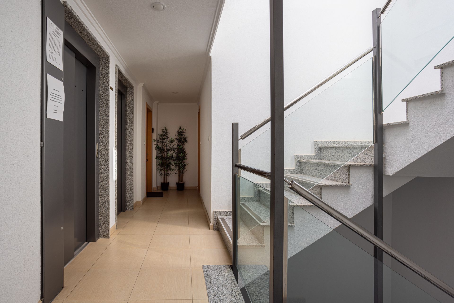 A hallway with two dark doors on the left, cream tile floors, potted plants at the end, and a modern glass-railed staircase.