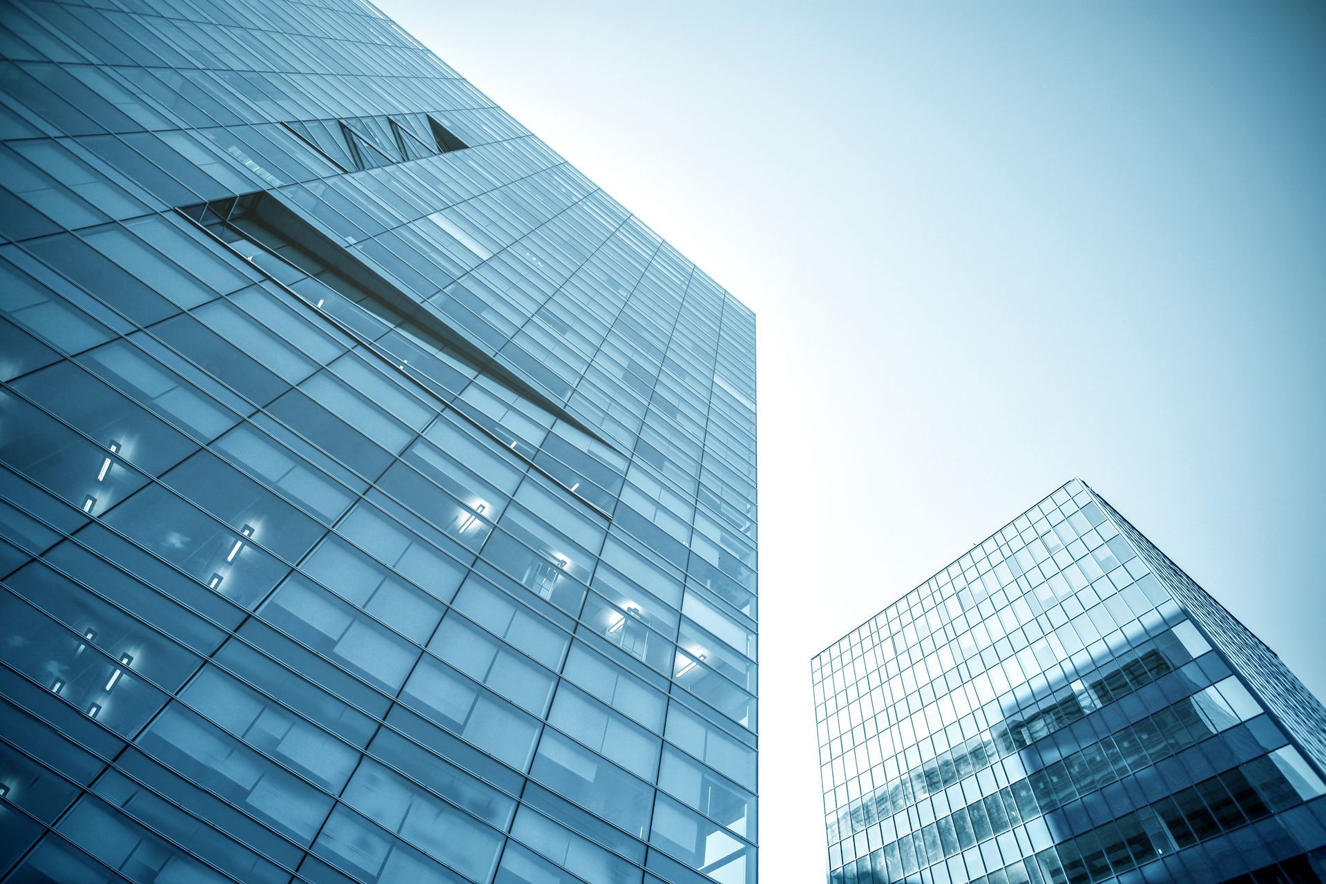 Low-angle view of two modern, blue-tinted glass skyscrapers against a bright sky.