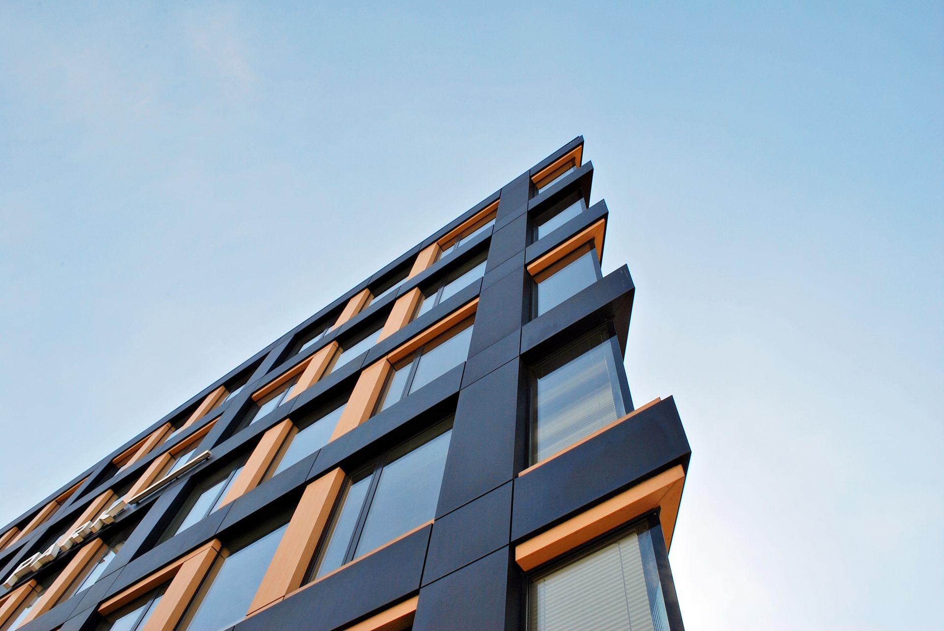 A modern building corner viewed from below against a blue sky, featuring dark panels and wood-toned window frames.