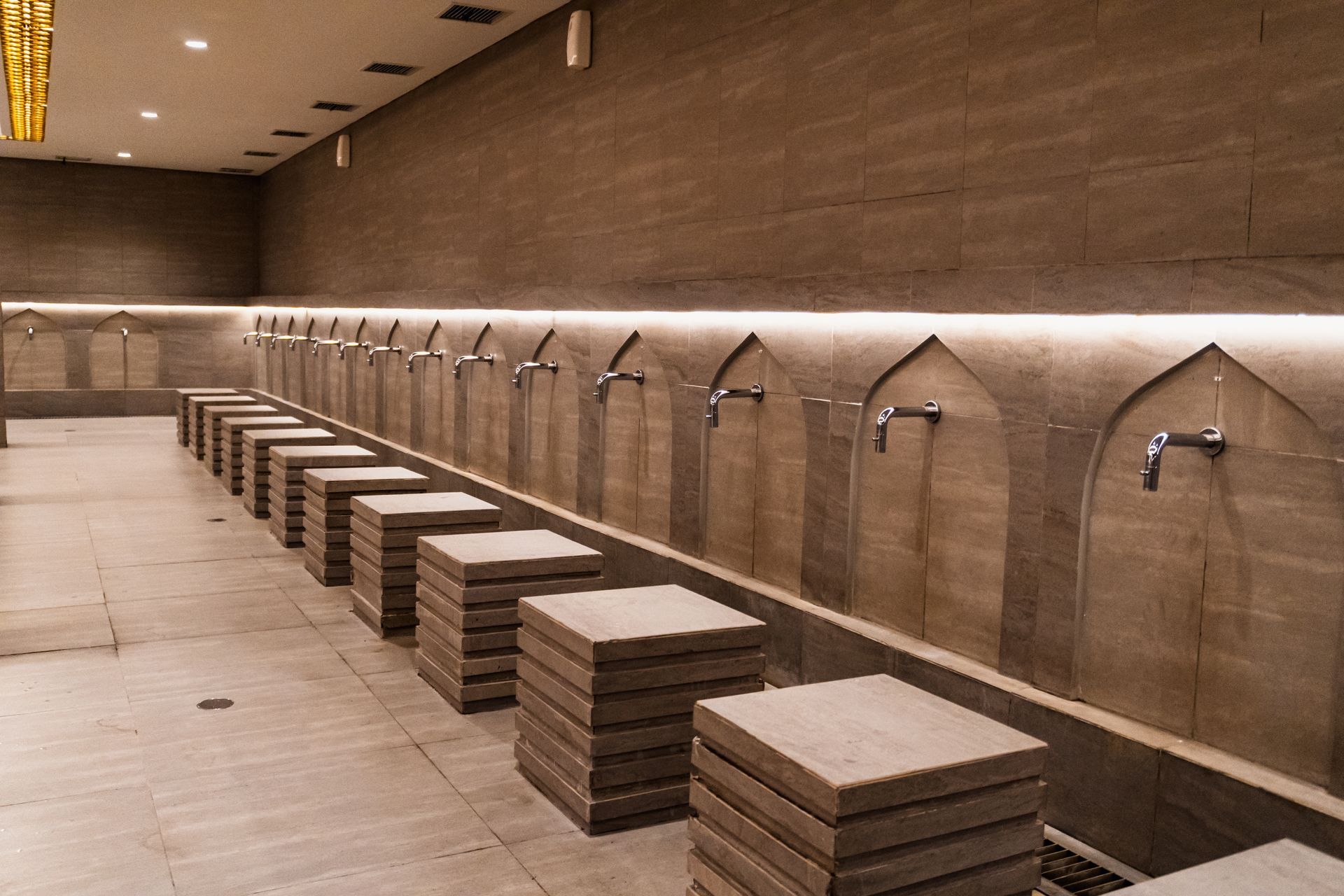 A row of faucets for ritual ablution with stone seats in a modern, tiled room with indirect lighting.
