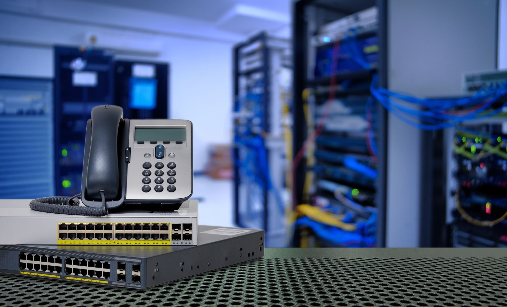 A VoIP phone sits on top of network switches in a server room, with equipment racks blurred in the background.